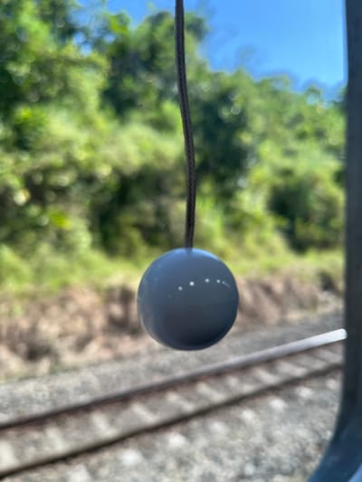 A grey, spherical object, likely a plumb bob, hangs from a dark string in the foreground, suspended in front of a blurred background of lush green foliage and a bright blue sky. The object has several small, white dots arranged in a pattern on its surface. Below the hanging object, out of focus, are two parallel train tracks running horizontally across the frame. The scene appears to be outdoors, possibly from a moving vehicle, given the blurred background and the presence of train tracks. The overall impression is one of movement and a fleeting glimpse of nature. There is no information about the location in Cilongok, Indonesia.