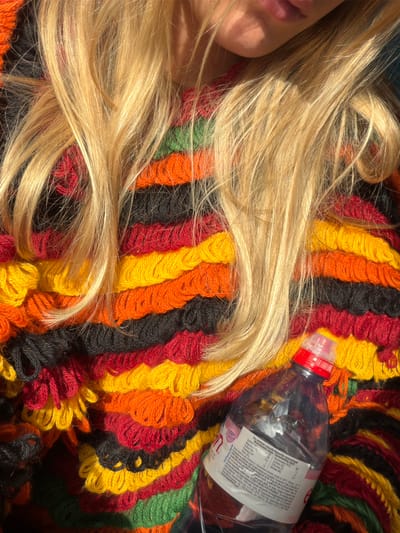 The scene, located in Mios, France, presents a close-up of an individual. The lower portion of a face, including lips, is visible in the upper left quadrant. A mass of light blond hair cascades across a distinctive garment. This garment features horizontal stripes of looped yarn in alternating colors including black, red, orange, yellow, and green. In the lower right quadrant, a clear plastic Evian water bottle with a red and clear sports cap is positioned on the colorful garment. The bottle contains a clear liquid and its white label displays text in French and Dutch. Direct sunlight illuminates the hair and parts of the woven fabric.
