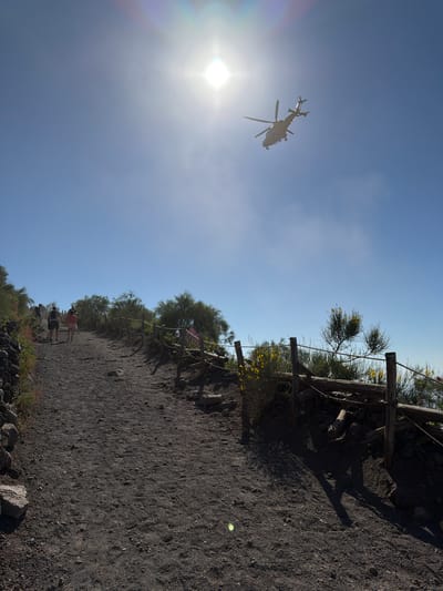 An eye-level, medium outdoor shot captures a dirt path winding uphill under a bright, clear blue sky. The path is lined with a rustic wooden fence on the right, made of posts and rope, separating it from sparse, dry vegetation with yellow flowers. On the left, the path is bordered by dark volcanic rocks and low-lying green and pink wildflowers. Several people are walking up the path, appearing as small figures in the distance. A helicopter flies in the upper right quadrant of the sky, its rotors visible. The sun is high and bright, creating lens flare and casting shadows on the path. The scene suggests a hiking trail, possibly on Mount Vesuvius, given the volcanic rock and barren landscape typical of the area near Ercolano, Italy.