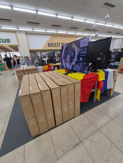 This image captures a retail environment, likely an electronics store in Käerjeng, Luxembourg. The primary subjects are several large, stacked cardboard boxes of Samsung Neo QLED televisions. The boxes are lined up on the floor, suggesting a display or recent delivery. Above the boxes, a large television screen is visible, displaying promotional content with text indicating "Neo QLED," "AI Gen2 Processor," and "AI TV."
In the background, more electronics, including televisions, are displayed on shelves. The store features bright fluorescent lighting from a tiled ceiling. Decorative soccer balls are suspended from the ceiling by strings, hinting at a sporting event promotion, possibly the World Cup or a European Championship. Flags of various nations, including Germany and Belgium, are draped over some of the electronics, further reinforcing the sporting theme.
Several people are visible in the background, appearing to be shoppers or employees. They are casually dressed and moving through the store. The overall atmosphere suggests a typical retail day, with products on display and customers browsing. The signage visible, such as "MULTI T" and "tus," points to the specific store or department. The time of day is not explicitly indicated, but the bright artificial lighting suggests it could be any time during store hours. The setting is indoors, with a tiled floor and typical retail store infrastructure. There are no overt emotional cues displayed by the people in the image.