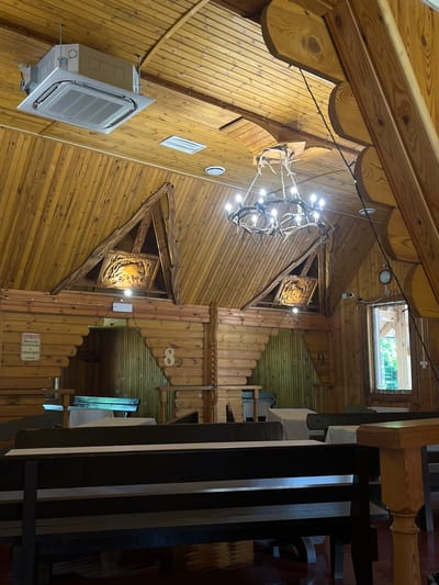The image shows the interior of a wooden building, possibly a restaurant or cafe, in the Шарашэўскі сельскі Савет, Belarus. The architecture is rustic, with exposed wooden beams and paneling. In the foreground, there are dark-colored wooden benches and tables. In the background, there are more wooden benches and tables, some covered with white tablecloths. A large, antler chandelier hangs from the ceiling, illuminating the space. There are also decorative carvings on the wooden walls and a window with a view of greenery outside. An air conditioning unit is visible on the ceiling. The scene appears to be empty of people.