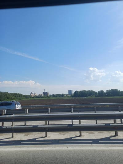 The image shows a view of a rural landscape with buildings and trees in the distance, taken from a highway. In the foreground, there are multiple levels of metal guardrails, with shadows cast on the ground below, suggesting a sunny day. A silver SUV is visible on the road behind the guardrails, driving away from the viewer. In the mid-ground, there are fields of dark soil and a line of dense green trees. Beyond the trees, several multi-story buildings and a construction crane are visible, indicating development. The sky is a clear blue with a few wispy white clouds. The scene is characteristic of the outskirts of a city, possibly on the outskirts of Krasnoobsk, Russia, given the presence of both agricultural land and developing urban areas.