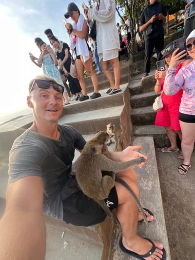 This image captures an outdoor scene at a tourist location on concrete steps in Bunga Mekar, Indonesia, during daylight hours. In the immediate foreground, a Caucasian male, with blonde hair and sunglasses resting on his head, wearing a grey t-shirt, black shorts, and black flip-flops, holds a camera device to take a selfie, smiling directly at the lens. A long-tailed macaque monkey is positioned on his left thigh, extending its right forelimb towards his open left hand. A second macaque is partially visible near his right leg. A third macaque sits on the concrete step to the man's right, observing the interaction.
Behind the foreground subject, a group of approximately ten individuals, identified as tourists, are visible on a series of concrete steps ascending into the background. Many are holding smartphones vertically or horizontally, seemingly recording or photographing the scene. Notable individuals include a woman in a dark baseball cap with a red can tucked into her shorts, a person in a white tunic, a woman in a light blue patterned dress, a woman in a red top and shorts, and another woman in a purple patterned long-sleeved top and black shorts wearing sunglasses. The background on the left is dominated by a bright, hazy sky, suggesting a low sun angle, while the right displays blurred green foliage. The environment consists of rough concrete steps and natural elements.