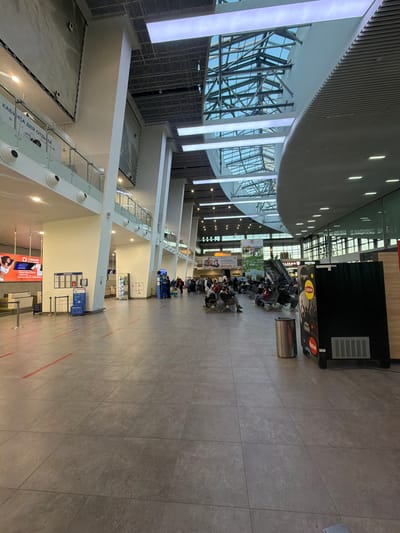 The image depicts an interior space in Bolshoye Savino, Russia, presumably at an airport or transportation hub, given the presence of seating areas with people waiting. The space is characterized by modern architecture, featuring a curved ceiling with exposed structural elements and large windows. There are rows of seats occupied by individuals, suggesting a waiting area or a queue for a service. There are also kiosks or vending machines. The floor is tiled.