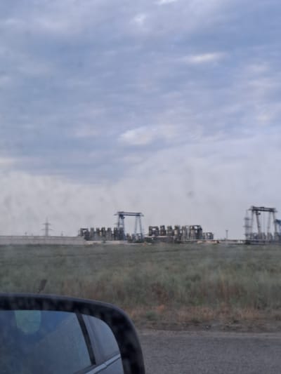 The image shows an industrial landscape with multiple structures resembling oil pumpjacks in the distance. The sky is overcast with gray clouds. In the foreground, a portion of a car's side mirror is visible, reflecting a blurred, blue-toned scene. A grassy field with some sparse vegetation stretches between the car and the industrial area. A power line tower can be seen to the left of the industrial complex. The overall scene suggests an industrial or oil field area in a rural or semi-rural setting. The location is identified as Batyr Rural District, Kazakhstan.