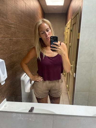 A woman with blonde hair takes a selfie in a restroom. She is wearing a maroon tank top and beige shorts. She has a ring on her left ring finger and a bracelet on her left wrist. Her left hand is on her hip. In the background, there is a brown tiled wall, a white hand dryer, and several doors. The woman is holding a black cell phone with a floral case in her right hand, facing the mirror. The sink is visible in the foreground, and the floor is tiled. The lighting appears to be artificial. The location is identified as a public restroom in the city of Agoy, Russia.