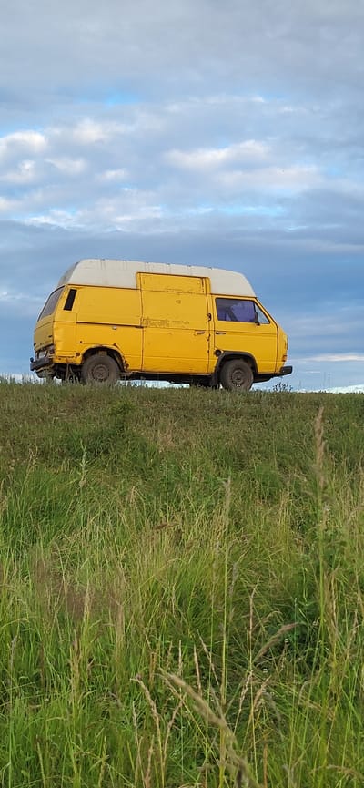 The image shows a yellow van parked on a grassy embankment under a cloudy sky. The van is old and appears to be in disrepair, with rust and peeling paint visible. It has a white roof extension and dark tinted windows. The foreground is dominated by tall, green grass with some dry stalks interspersed. The background consists of a cloudy sky with patches of blue visible. The scene is devoid of any people or human activity. The location is identified as Hetmanivka, Ukraine.