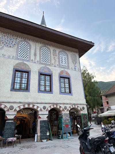 This is a photograph of the Colored Mosque in Travnik, Bosnia and Herzegovina. The mosque is a historical building known for its colorful exterior decorations. In the foreground, there are several motorcycles parked on a cobblestone street. The building itself features arched entrances on the ground floor, with shops or stalls set up behind them. The upper floors have distinctive windows and intricate decorative patterns. A young girl is visible in the background, walking on a street that leads to other buildings and a shaded area with seating. The sky is partly cloudy, and a green hillside is visible behind the buildings.