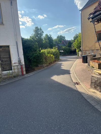 The image depicts a street scene in Hüffler, Germany, on a sunny day with scattered clouds. The perspective is from street level, looking down a paved road that curves to the right.
On the left side of the street, a white building with a weathered wooden door is partially visible. A gray downspout runs along the side of this building, with a red pipe section near the bottom. The building has a textured facade and a checkered tile pattern at its base.
The right side of the street features a yellow building with a tiled roof and a stone-clad lower section. Two dark-colored trash bins are situated near the yellow building. A paved walkway with red brick pavers runs alongside the road. A concrete planter box filled with dry vegetation is also present.
Mature green trees and dense hedges line both sides of the street, creating a suburban or village atmosphere. In the background, several houses with red-tiled roofs are visible, nestled among the trees. Road signs are faintly discernible in the distance. The road surface is asphalt, and a storm drain grate is visible on the right side. The sunlight casts long shadows across the road.