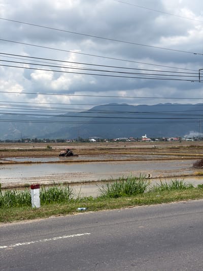 This image depicts a rural landscape in Diên Lạc, Vietnam, likely a rice paddy field. In the foreground, a paved road with a white line runs horizontally across the bottom of the frame, bordered by a grassy verge. A white and red marker post stands on the verge. Beyond the road, a flooded agricultural field stretches towards a distant treeline and mountains under a cloudy sky. A red tractor is actively plowing the muddy field, churning up water and soil. Several other figures appear to be working in the field at a distance. In the background, a village with various buildings, including a white-domed structure possibly a mosque or temple, is visible against the backdrop of rolling hills and mountains. Power lines are strung across the sky, intersecting the scene.