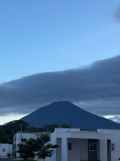 The image depicts the Volcano of San Miguel (Chaparrastique) as seen from the city of San Miguel, El Salvador, during dusk or early morning. The volcano, a prominent, dark blue conical shape, dominates the background, partially obscured by a large, dark grey cloud formation stretching across the upper portion of the frame. The sky above the clouds transitions from a lighter blue to a deeper hue. In the foreground, modern, white, two-story houses with minimalist architectural designs are visible, set against a backdrop of lush green trees. One house features a dark grey facade around the entrance and a "SE VENDE" (For Sale) sign in a window. Power lines are also visible stretching horizontally across the scene, in front of the volcano and houses. The lighting suggests low light conditions, characteristic of twilight. No people or active events are depicted.
