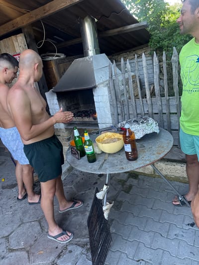 In Varbak, Bulgaria, three men are gathered around a rustic outdoor cooking area. A brick oven with a metal chimney is central to the scene, with a fire burning inside. Two men, shirtless and wearing shorts, stand near the oven. One, bald, is positioned to the left, while the other, wearing dark green shorts, is in the foreground. A third man, wearing a bright green shirt and light blue shorts, stands to the right.
A circular metal table is set up in front of the oven, laden with items. Two green glass beer bottles are prominent, along with a brown glass bottle. A yellow bowl filled with food, possibly a salad or dip, sits on the table, next to a fork. A portion of food wrapped in aluminum foil is also visible. The ground is paved with interlocking gray stones, and a metal grill sits on the lower right side of the image. The setting appears to be a backyard or a communal outdoor space, with a wooden fence and greenery in the background.