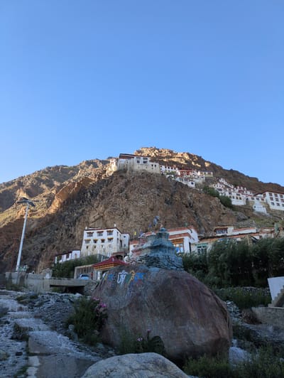 The image shows a monastery complex built on a rocky mountainside in Yulang, India. The monastery buildings are white with red-trimmed roofs and windows. They are spread across the hillside, some appearing to be several stories high. In the foreground, there is a large boulder with colorful Tibetan Buddhist inscriptions painted on it. A small stupa, also in the Tibetan Buddhist style, sits atop the boulder. A stream flows over rocks in the lower left of the frame. A street lamp is visible on the left side of the image. The sky is clear and blue.