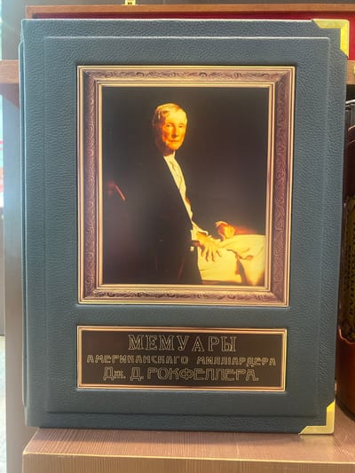 The image shows a dark teal bound book with a portrait of an elderly white man in a suit inside a decorative gold frame. Below the portrait is a brass plaque with text in Russian. The book is resting on a wooden surface, likely a shelf, in what appears to be an indoor setting. The overall presentation suggests it is a collection or a special edition of a book.