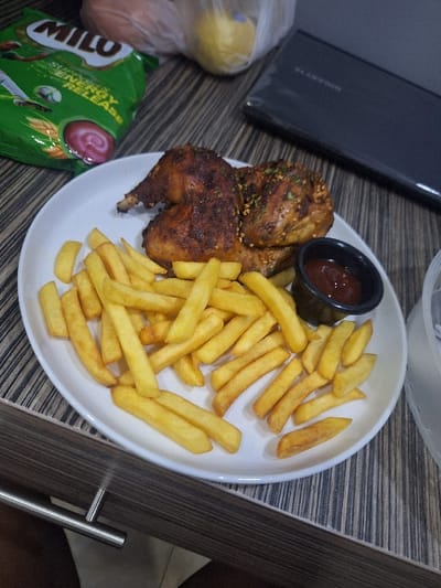 A white plate holds a meal of golden French fries and roasted chicken. A small black container filled with red sauce sits beside the chicken. A green Milo packet is partially visible, along with a plastic bag containing an orange fruit, possibly an orange, and a lemon. A black tablet or laptop rests near the plate. The meal appears to be set on a table with a patterned surface. A chrome handle is visible on the side of the image.