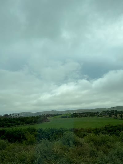 This media file depicts an overcast, possibly rainy, landscape in Fanmore, Ireland. The image is oriented vertically, with the sky occupying the left side and a rural scene on the right.
The sky is filled with thick, gray clouds, suggesting inclement weather. The landscape below consists of rolling green hills and fields, bordered by dense green trees and foliage. In the distance, the outlines of mountains can be seen, shrouded in the mist or clouds. There are hints of human presence, including what appear to be utility poles and possibly some buildings nestled amongst the trees. On the far right, there is a glimpse of what could be a quarry or industrial site with heavy machinery, including a crane and a truck.
The overall mood is somber, consistent with the cloudy and potentially wet weather. The time of day is indeterminate due to the lack of direct sunlight. The location is identified as Fanmore, Ireland. There is no visible text in the image.