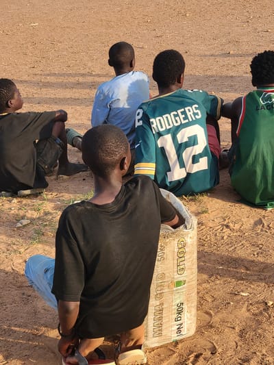 The image depicts a group of five young men outdoors, gathered around a sack labelled "GOLD 50kg Net" and "MAMA GOLD 50kg". They are located on bare, reddish-brown earth, possibly in a rural or undeveloped area. The men are dressed in casual clothing, including jerseys and t-shirts. One wears a jersey with "RODGERS 12" printed on it. The focus of the scene is the sack, around which the group is gathered. The setting appears to be sunny, based on the brightness and shadows. The location is stated as Ibo, Nigeria.