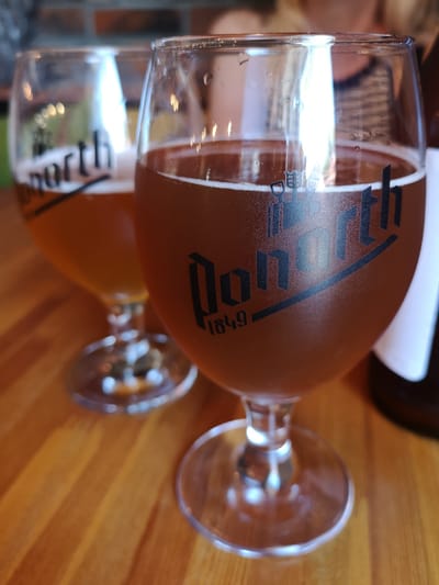Two stemmed glasses of amber-colored beer are on a wooden table. The glass in the foreground has a logo that reads "Donnorth 1849". The glass in the background has a logo that reads "north". The background is blurred, showing a brick wall and a person wearing a striped shirt. There is no indication of the location being Yantarny, Russia.