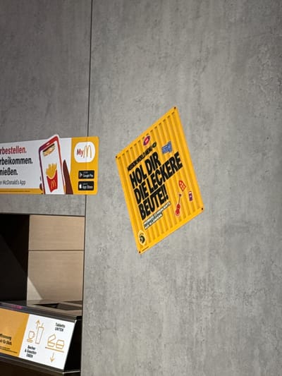 The image shows a section of a concrete wall with various stickers and signage. On the left, a white and orange sticker advertises the "MyMcDonald's App" with a depiction of a phone displaying a burger and fries. Below this sticker, a portion of a shelf unit is visible. To the right of the MyMcDonald's sticker, a bright yellow square sticker with bold black text reads "HOL DIR DIE LECKERE BEUTE!!". This sticker features various smaller icons and the text "PROBIERE 23 HIGHLIGHTS AUS 16 LÄNDERN". The concrete wall has a textured, grey appearance. In the lower left corner, another sign with orange and white elements displays icons related to trash disposal, indicating "Becher & Geschirr OBEN" (cups and cutlery above) and "Tabletts UNTEN" (trays below). The presence of the McDonald's branding indicates this scene is likely within a McDonald's restaurant, possibly in Saalfeld/Saale, Germany, given the German language on the signage. There are no people visible in the image.