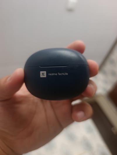 A dark blue, oval-shaped charging case for wireless earbuds is held in a person's hand. The case has a small, white logo and text that reads "realme TechLife" on its surface. The background is blurry and appears to be an indoor setting. This image was taken in Denov, Uzbekistan.