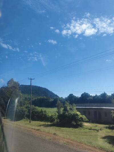 The image shows a rural scene in Kibirichia ward, Kenya. The foreground consists of a paved road with a dirt shoulder and green grass. A concrete building with a corrugated metal roof is present on the right side of the frame, partially obscured by trees and vegetation. Behind the building is a field and a forested hill. A power pole with multiple wires is visible, spanning the sky above the landscape. The sky is bright blue with scattered white clouds. The image appears to be taken from inside a moving vehicle, with a partial view of the vehicle's interior on the left.