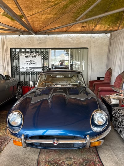 The image shows a blue vintage car parked in front of the "Wijnland Auto Museum - Leslie Boshoff" in Kraaifontein, South Africa. The museum's sign is visible through the metal security gate. Other cars are partially visible in the foreground. Part of a tent structure is seen at the left side of the frame. Some antique red chairs and other objects are visible behind the car on the right side. An ornate rug lies on the ground next to the car.