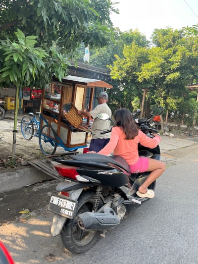 In the city of Sugih Waras, Indonesia, a young girl with long dark hair, wearing a coral pink long-sleeved shirt and pink shorts, is perched on the back of a black scooter. Her legs are bent, and she is wearing beige clogs. The scooter's license plate reads "W 3183 R" and "09-31." Beside the scooter, a man in a beige t-shirt, dark shorts, and a grey baseball cap stands by a wooden food cart. He is holding a woven fan and appears to be attending to the cart, which has a sign in Indonesian that reads "SATE AYAM" (Chicken Satay) and "CAK UMAR." In front of the food cart is a blue bicycle with two large wheels. The scene is set outdoors on a paved surface, with lush green trees and foliage surrounding the area. The sunlight casts shadows on the ground, suggesting it is daytime.