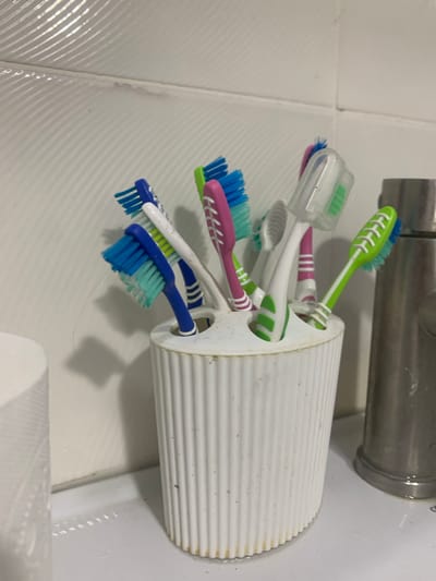 A close-up, eye-level shot displays a white toothbrush holder filled with several toothbrushes. The toothbrushes are of various colors, including blue, green, pink, and white. Some of the toothbrushes have protective caps. The holder is ribbed and appears to be made of plastic. Behind the toothbrush holder, a white tiled wall with horizontal grooving is visible. To the right of the holder, a portion of a metallic faucet is present. The scene is set on a white countertop, which shows signs of minor water residue and some dirt. A thin, light-colored strand, possibly a hair, is visible hanging from one of the toothbrushes. The overall impression is that of a bathroom or a similar hygiene-focused area. The location is identified as Glodeni, Moldova.