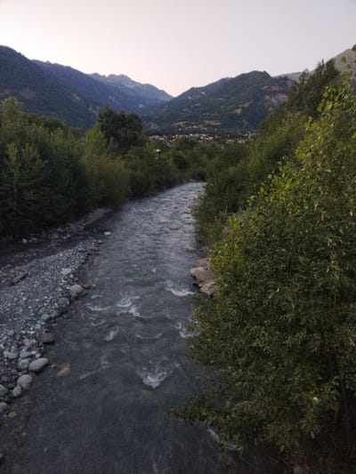 This image depicts a tranquil natural scene in the vicinity of Saint-Étienne-de-Cuines, France, likely captured at dusk. A wide, fast-flowing river with turbulent water occupies the center of the frame, its surface showing whitecaps and ripples. The riverbanks are lined with lush green trees and dense foliage. On the left bank, a stretch of gray gravel and scattered rocks is visible. In the background, rolling hills and mountains rise, covered in vegetation and dotted with a small village, its lights faintly visible. The sky above is a pale, hazy gradient, suggesting the fading light of day. No people or distinct structures are identifiable in the foreground or midground.