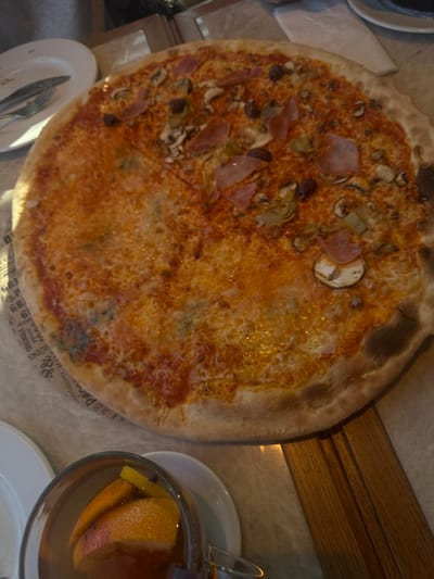 A large, round pizza occupies the center-right of the frame, resting on a light-colored paper liner printed with partial text including "GRILL" and "Palace." The pizza exhibits varied toppings, suggesting a multi-section composition. One prominent section contains sliced mushrooms, folded slices of light pink ham, black olives, and green artichoke hearts. Another significant section features melted cheese over a red sauce base, with visible areas of blue cheese intermingled. The crust is uniformly golden-brown. To the upper left, a white ceramic plate holds a fork and knife. In the lower-left, a clear glass mug contains a reddish-brown beverage with slices of grapefruit and lemon, along with small dark spices, positioned on a small white saucer. A wooden surface is partially visible on the right side of the table. The scene is illuminated by overhead light, casting reflections on the table surface. The setting is an indoor dining area in Mysiadło, Poland. No individuals are visible.