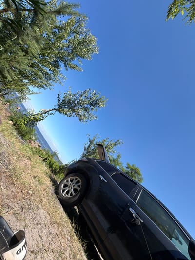 The image depicts a black SUV parked on a dirt and grass embankment next to a body of water, likely a lake or river. The car's rear hatch is open. The scene is set under a clear blue sky. Trees with green foliage are visible in the foreground and background, some overhanging the water. The water appears calm, with a hint of a distant shoreline or landmass. In the lower left corner of the frame, a portion of a black and white slide sandal with the word "GIVEN" is visible. The photo appears to be taken from a low angle, with the camera tilted. The location is specified as Kozarovychi, Ukraine.