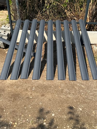 In Maļutki, Latvia, a row of eight dark gray, ribbed metal fence pickets are arranged vertically on a dirt and gravel surface. The pickets are leaning against a dark metal fence structure and a concrete foundation. To the left of the pickets, several concrete blocks are stacked. The background consists of dry, brown vegetation. The lighting suggests it is daytime, with shadows cast by the pickets.