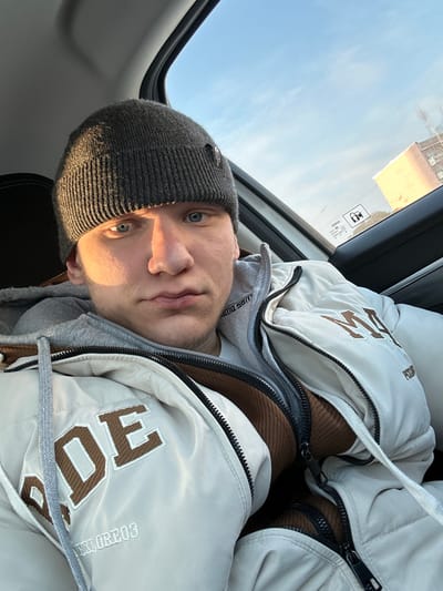 A young adult male is depicted from the chest up, seated inside a vehicle, likely in the front passenger position. He has light skin, blue eyes, and short facial hair, and is looking directly forward. He wears a dark grey ribbed knit beanie with a small dark patch on the left side, a light grey hooded sweatshirt, and an unzipped light beige puffer jacket. The jacket displays large brown block letters, including "RDE" on the left chest and "M" on the right chest, along with smaller text "PPLORE03" and "PS." A brown textured inner layer is visible beneath the jacket. Strong sunlight illuminates the right side of his face. Through the vehicle's right-side window, a clear blue sky with minimal clouds is visible above a distant, multi-story, light-colored building, possibly residential or commercial. The interior of the vehicle features dark trim, and a warning label is affixed to the window glass. This scene is located in the city of Dobryanka, Russia.