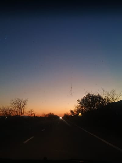 The image shows a view from a vehicle moving along a road at dusk or dawn. The sky transitions from blue on the left to orange on the right, indicating either sunrise or sunset. The road is on the right side of the image, with roadside vegetation including bare trees and possibly bushes on either side. A traffic sign can be seen along the side of the road. A light is also seen. The specific location cannot be determined as Zimnitsa, Bulgaria as the image does not contain identifiable features or objects unique to that specific place.