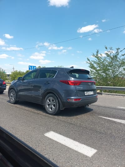 A dark gray Kia Sportage is parked on a paved road in Krymsk, Russia, on a bright, clear day with a blue sky and scattered white clouds. The car is facing to the left, with its rear visible to the viewer. The license plate reads "M852AY 799". A metal guardrail runs along the right side of the road, and lush green trees are visible behind it. In the distance, buildings and power lines can be seen. A road sign with arrows indicating lanes for straight and left turns is visible in the background. The road surface is asphalt with white lane markings.