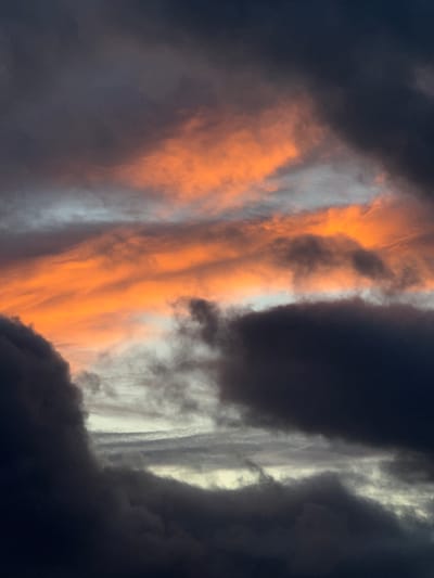 The image captures a dramatic and dynamic sky, likely during sunrise or sunset over Fécamp, France. The scene is dominated by an impressive array of clouds, showcasing a striking contrast between vivid light and profound darkness.
A prominent central band of clouds glows intensely with rich orange and amber hues, illuminated by the low-angled sun. These brightly lit clouds appear as swirling, elongated forms, stretching vertically across the frame, with some lighter white and pale gray sections interspersed within the vibrant orange. This luminous core is sharply bordered on both sides by heavy, dark gray to deep blue-black clouds, suggesting dense storm formations or deep shadows.
The textures of the clouds vary significantly; the orange-lit areas appear softer and more diffused, while the darker masses possess a more turbulent, dense, and opaque quality. The interplay of light and shadow creates a powerful sense of depth and movement in the sky. No specific objects, people, or discernible text are visible in the frame. The overall atmosphere is one of natural grandeur and dramatic beauty, hinting at powerful weather phenomena underscored by the fleeting beauty of dawn or dusk.