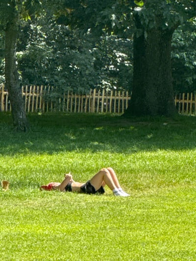 The image shows a person lying on a grassy lawn. The person is wearing white socks, black shorts, and a black top. Their legs are bent at the knees, with one foot resting on the ground and the other extended upwards. A small red object is held in one hand. The setting appears to be outdoors, with a large tree and a wooden fence visible in the background. The grass is green and well-maintained, suggesting a park or garden. The lighting indicates it is daytime, with shadows cast by the tree. The overall impression is one of relaxation or perhaps light exercise. There is no visible text in the image.