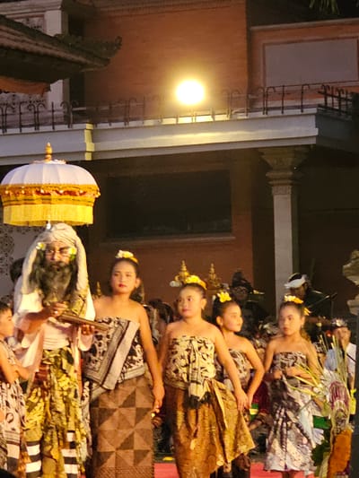 This media file captures a vibrant performance, likely a traditional Balinese dance, taking place outdoors at night, possibly in Denpasar, Indonesia. The scene is illuminated by a bright, warm light, casting a soft glow on the elaborately dressed performers and intricate surroundings.

The primary subjects are several young women, adorned in traditional costumes featuring patterned fabrics and floral headpieces. They appear to be dancers, their poses suggesting graceful movement and storytelling. One performer, positioned in the foreground, is more prominently featured, with a white and gold conical headdress, possibly a ceremonial object, placed beside her. Another figure, seemingly an older man with dark, long hair and a white head covering, is partially visible, his face expressing a serious or perhaps theatrical demeanor. The performers' attire is rich with detail, incorporating various textiles and ornamentation.

The setting seems to be an outdoor stage or courtyard with architectural elements that hint at a traditional Indonesian structure. A warm, bright light source, possibly a large lamp, is visible on the left side of the frame, illuminating a portion of the backdrop which appears to be a building with textured walls and decorative railings. The overall atmosphere is one of cultural celebration and artistic expression, with the night setting adding to the mystique and drama of the performance. The expressions on the faces of the performers, particularly the young women, convey a sense of focus and poise.