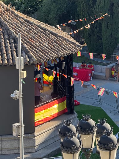 The image shows an outdoor scene with Spanish flags decorating the area. In the foreground, there are ornate street lamps with crowns on top. Behind the lamps, a large black wall is visible, with a metal pole attached to it. On the pole, there are electrical boxes and a security camera. Further back, a tiled roof structure with a weathered appearance is prominent, and a man in a pink shirt is standing near a stage adorned with Spanish flags. A red table with a logo and some text is set up, with two individuals sitting behind it. In the background, more Spanish flags are strung across wires, and trees and a stone bench are visible. The overall impression is of a festive or event-related gathering in an urban setting, likely Las Rozas de Madrid, Spain, given the prominent display of Spanish flags.