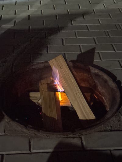 A medium shot captures a fire pit on a brick patio at night. Two pieces of firewood, arranged in an L-shape, are burning in the center of the metal fire pit. Flames, predominantly yellow and blue, lick at the wood. The surrounding brickwork is visible, with shadows cast from an unseen light source. The scene is set in Kurkut, Russia.