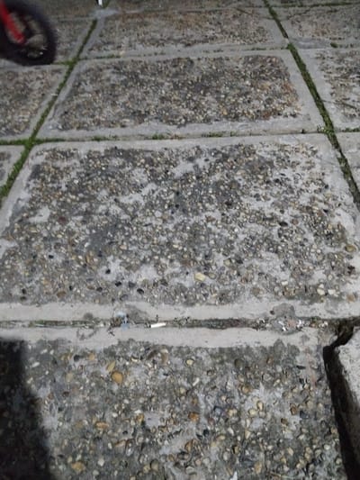 This media file captures a close-up, low-angle view of a pathway made of square concrete pavers embedded with pebbles. The pavers are arranged in a grid pattern, with thin strips of grass growing in the narrow gaps between them. The surface of the pavers is textured and uneven due to the embedded stones, and some areas appear damp, suggesting recent rain or dew. Litter, including a discarded cigarette butt, is visible in the crevices between some of the pavers. In the upper left corner of the image, a portion of a black bicycle wheel with a red rim is visible, indicating the presence of a bicycle. The overall lighting suggests it may be dusk or dawn, or an overcast day, as there are no strong shadows. The scene is likely an outdoor walkway or patio. The location context provided is Palangka Raya, Indonesia. There is no visible text in the image.