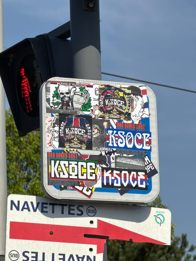 The image shows a close-up of a street sign covered in stickers, located in Rungis, France. The sign is a white rectangular board with blue lettering that reads "NAVETTES (BUS)" and a red arrow pointing to the right. Below this, another sign with similar lettering is partially visible.
On top of the "NAVETTES" sign, a smaller white board is affixed, also covered with various stickers. These stickers depict skulls, trophies, text such as "FINALE MUNICH", "BAD GONES 1987", and "K-SOCE", and some feature camouflage patterns. A pedestrian traffic light with a red illuminated figure is visible to the left of the signs. The background consists of a blue sky with some clouds and greenery. The overall scene suggests an urban or semi-urban outdoor environment.