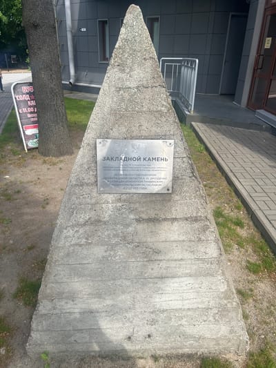 A concrete pyramid-shaped monument stands on a paved area in front of a modern building with grey tiled walls and a glass door. A metal plaque is affixed to the monument. A tree trunk is to the left of the monument. A sidewalk with brick-like paving extends from the foreground to the right of the image. The monument has a plaque that reads "ЗАКЛАДНОЙ КАМЕНЬ" (Founding Stone) and lists the location as "НА МЕСТЕ СТРОИТЕЛЬСТВА ЭКСПОЗИЦИОННО-ВЫСТАВОЧНОГО МУЗЕЯ-ЗАПОВЕДНИКА «ПРОРЫВ» БЛОКАДЫ ЛЕНИНГРАДА" (At the site of construction of the exhibition-museum complex "Breakthrough" of the Siege of Leningrad). It also states it was installed by Governor A. Yu. Drozdenko of Leningrad Oblast and consecrated by Bishop Tikhvin and Ludinopolislav Mstislav on May 8, 2015. This monument is located in Shlisselburg, Russia, specifically at the site of the "Breakthrough" Museum.