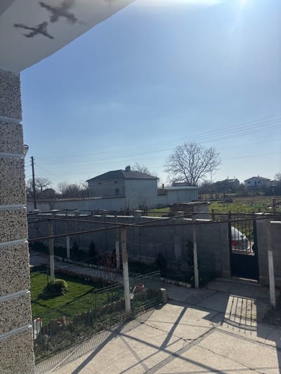 The image depicts an outdoor residential scene in Kalitinovo, Bulgaria, captured from an elevated vantage point on a clear, sunny day. The foreground on the left features a textured, light-colored concrete pillar. The lower foreground consists of a cracked concrete patio surface, marked by strong diagonal shadows cast from an unseen overhead structure. A green metal fence separates this patio from a small garden area with green grass, small plants, a red container, and a plastic bottle with a red cap.
Across the midground, a trellis system constructed from rusty metal poles and crossbars spans horizontally. Behind this, a grey concrete block wall forms a property boundary. To the right, a black metal gate is partially ajar, revealing the front section of a light-colored sedan parked within the property.
The background features several residential buildings. A prominent two-story house with a grey tiled roof and light-colored walls stands in the center-left. Additional houses are scattered across the horizon, including a white structure with a reddish roof on the far right. Leafless deciduous trees are visible, notably a large, prominent tree in the center-right. Overhead utility poles and multiple power lines traverse the upper midground. The sky is clear blue with bright sunlight, causing a lens flare in the upper right. Two dark, irregular smudges are present on the upper-left edge of the frame. No individuals are visible.