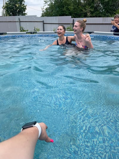 In the city of Plodopitomnik, Russia, two women are joyfully splashing in an outdoor swimming pool. One woman, with dark hair pulled back, wears a black swimsuit and is laughing. The other, with blonde hair styled in a bun, wears a colorful strapless bikini top and has her arm around the first woman. Behind them, another woman is visible in the background, holding a phone and appearing to take a photo. The pool water is a vibrant blue, with ripples and reflections from the overcast sky and surrounding trees. A fence and greenery are visible beyond the pool. In the foreground, a person's arm wearing a smartwatch and with bright pink fingernails is reaching into the water.