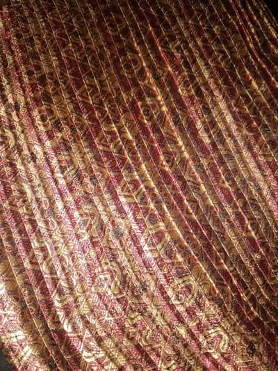 The image displays a close-up view of a pleated textile, observed in the city of Na Sang, Laos. The fabric features an intricate, repeating pattern predominantly in metallic gold and deep red (maroon) tones, with subtle darker accents. The material is uniformly pleated, forming distinct parallel ridges and valleys that extend diagonally across the entire frame, from the upper-left to the lower-right. The pleats exhibit varying degrees of light reflection, highlighting the shimmering quality of the gold threads and casting subtle shadows. No individuals, other objects, or discernible background environment are visible within the frame.