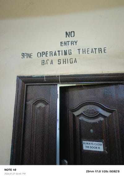 The image displays an interior scene in Gwammaja, Nigeria, focusing on a doorway within a medical facility. A light-colored wall features stenciled black text: "NO ENTRY", "SPINE OPERATING THEATRE", and "BA'A SHIGA" (Hausa for "No Entry"). Below this, a set of dark-paneled double doors is partially open, revealing a brightly lit space beyond. A white paper sign, affixed to the right door, displays black text reading "ALWAYS CLOSE THE DOOR !!!". No individuals are visible in the scene.