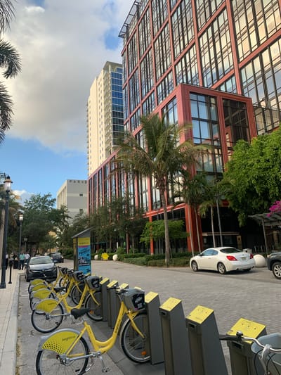 The image shows an urban scene in West Palm Beach, Florida, United States.
A row of yellow "brightbike" rental bicycles is parked on a paved sidewalk. Behind the bicycles, a white car is parked along the street. The "brightbike" logo and branding are visible on the bicycles and their docking stations.
Buildings of varying heights and architectural styles surround the scene. One building is characterized by a grid-like facade with a reddish-brown frame. Another building is a tall, white, high-rise structure. There are trees, including palm trees, dotting the landscape between the buildings.
Signage is visible indicating "PARKI", "CENTER OF Paradise", and "MORE TO EXPLORE" alongside the "brightbike" advertising, suggesting a tourist-oriented area. The sky is partially cloudy, with patches of blue visible.