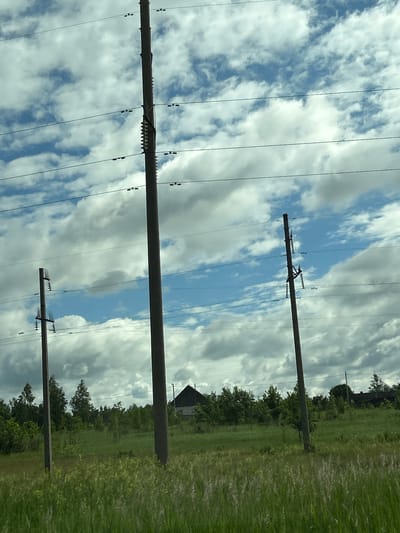 This is a medium-angle, eye-level outdoor shot taken from a moving vehicle. The scene depicts a rural landscape with three prominent utility poles supporting power lines against a sky filled with scattered clouds. The foreground is dominated by tall, green grass, some of which is blurred due to motion. In the midground, there is a dense line of trees, with a dark, triangular-roofed building partially visible behind them on the right side of the frame. The overall impression is one of a quiet, somewhat overcast day in the Belarusian countryside, likely within the Малевіцкі сельскі Савет area, given the characteristic wooden poles and rustic architecture. There are no people visible in the image.
