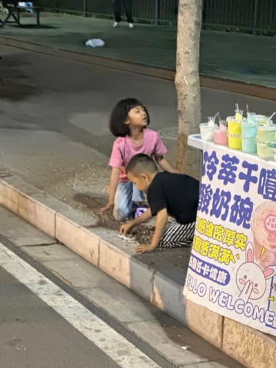 Two children, a girl in a pink shirt and a boy in a black shirt, are kneeling on the ground near a street vendor's cart in Eshan Yi Autonomous County, China. The vendor's cart displays various colorful drinks in clear plastic cups, along with signage written in Chinese characters and English. The girl looks upwards, her gaze directed towards the sky, while the boy is focused on something on the ground in front of him, holding what appears to be a small object. The ground around them is littered with small debris. A tree trunk is visible to the right of the children, and a paved sidewalk with a white stripe runs in the foreground. In the background, out-of-focus figures are visible, suggesting a public area with pedestrian traffic. The overall scene appears to be an outdoor setting, possibly a street or park, during the evening or late afternoon, as indicated by the lighting.